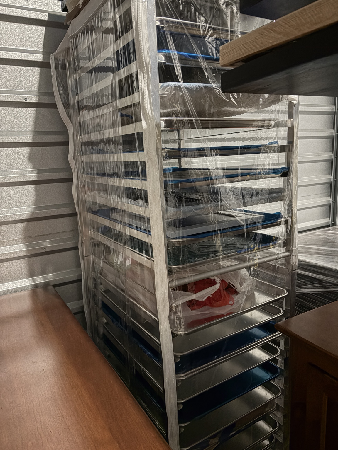 Commercial bakery sheet pan rack wrapped in a clear cover inside a storage unit next to wood furniture